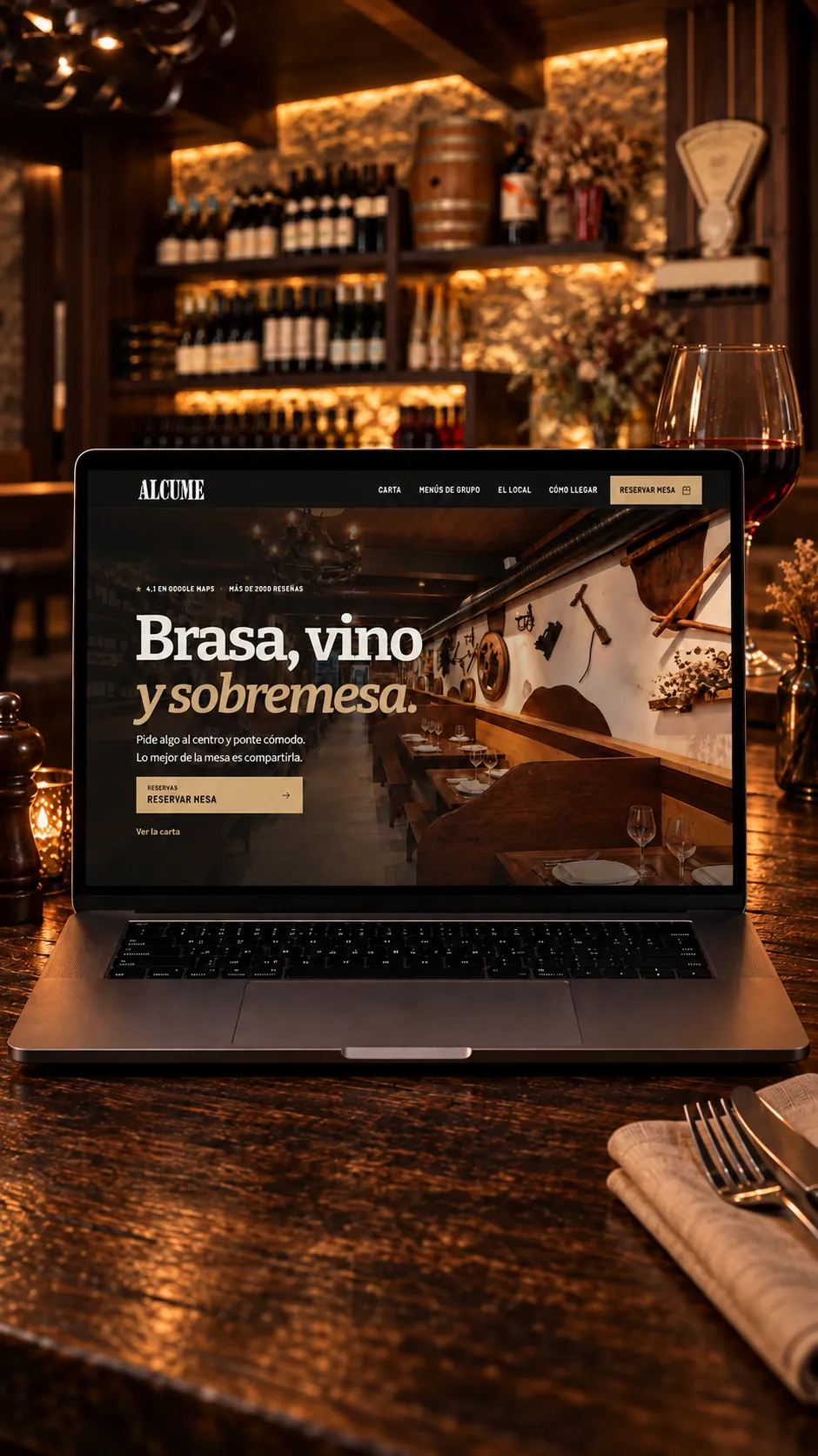 Presentación de la web de Alcume en un portátil, con una copa de vino y la bodega de piedra y madera al fondo.